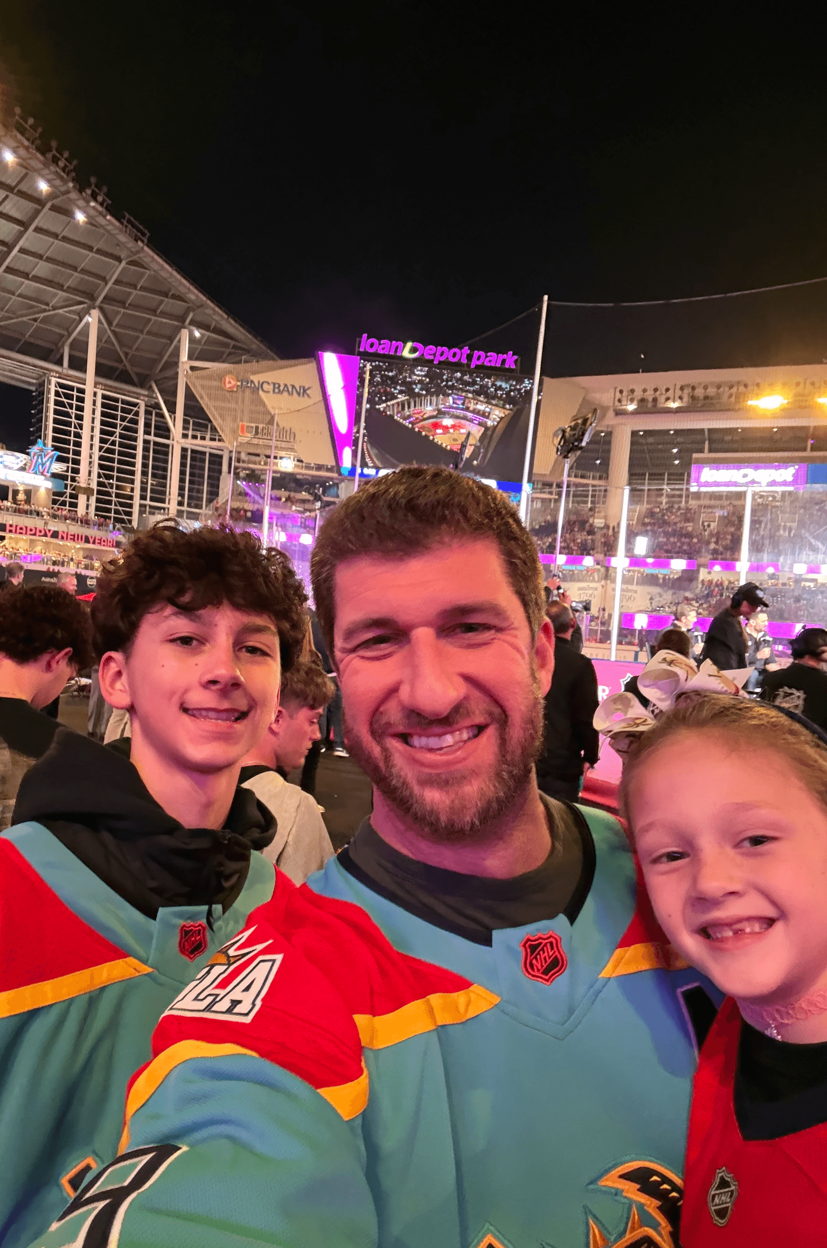 Three people smiling at a sports event, with a stadium and bright lights in the background.