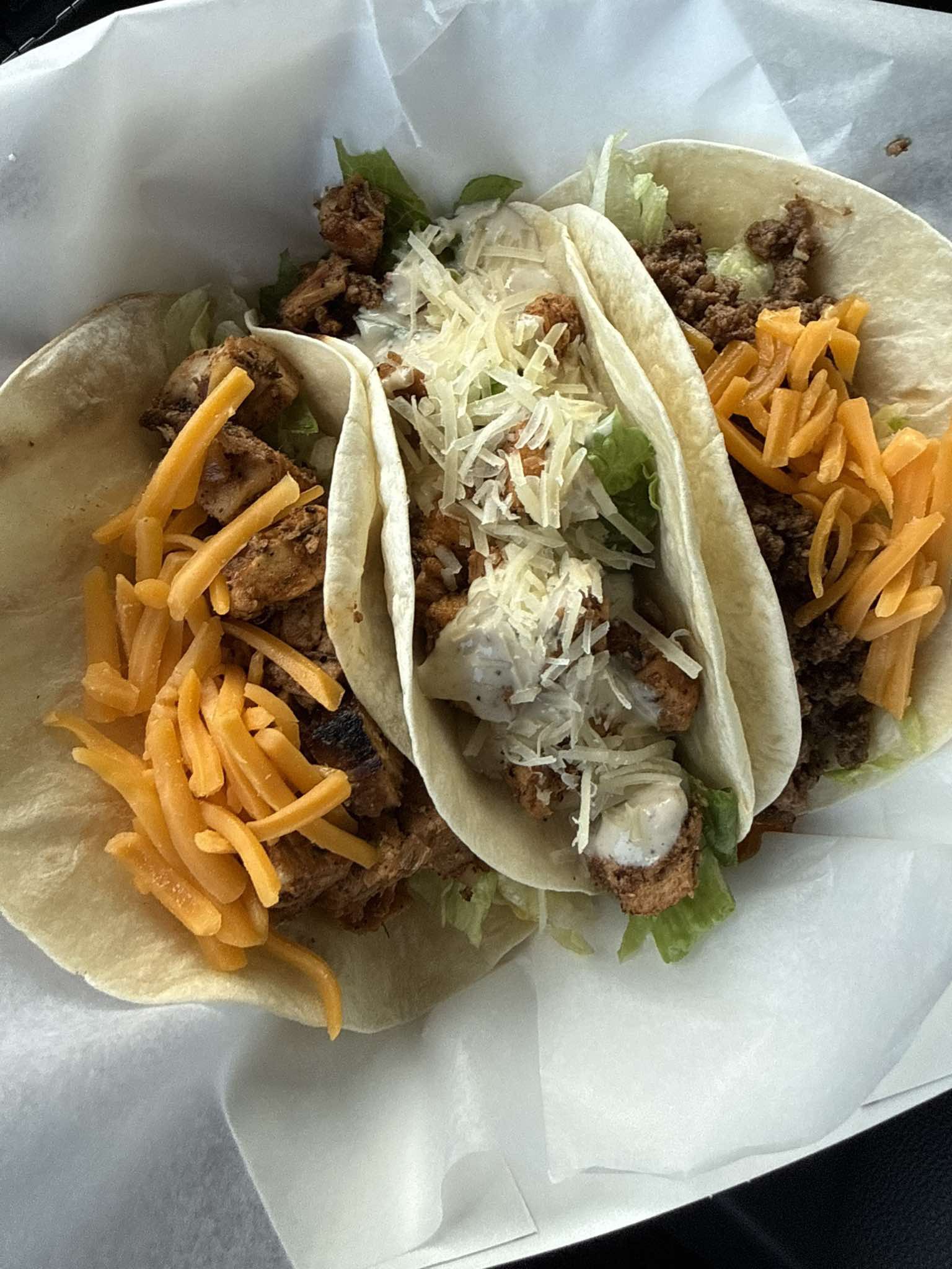 Three tacos with shredded cheese, lettuce, and seasoned meat in soft tortillas on white paper