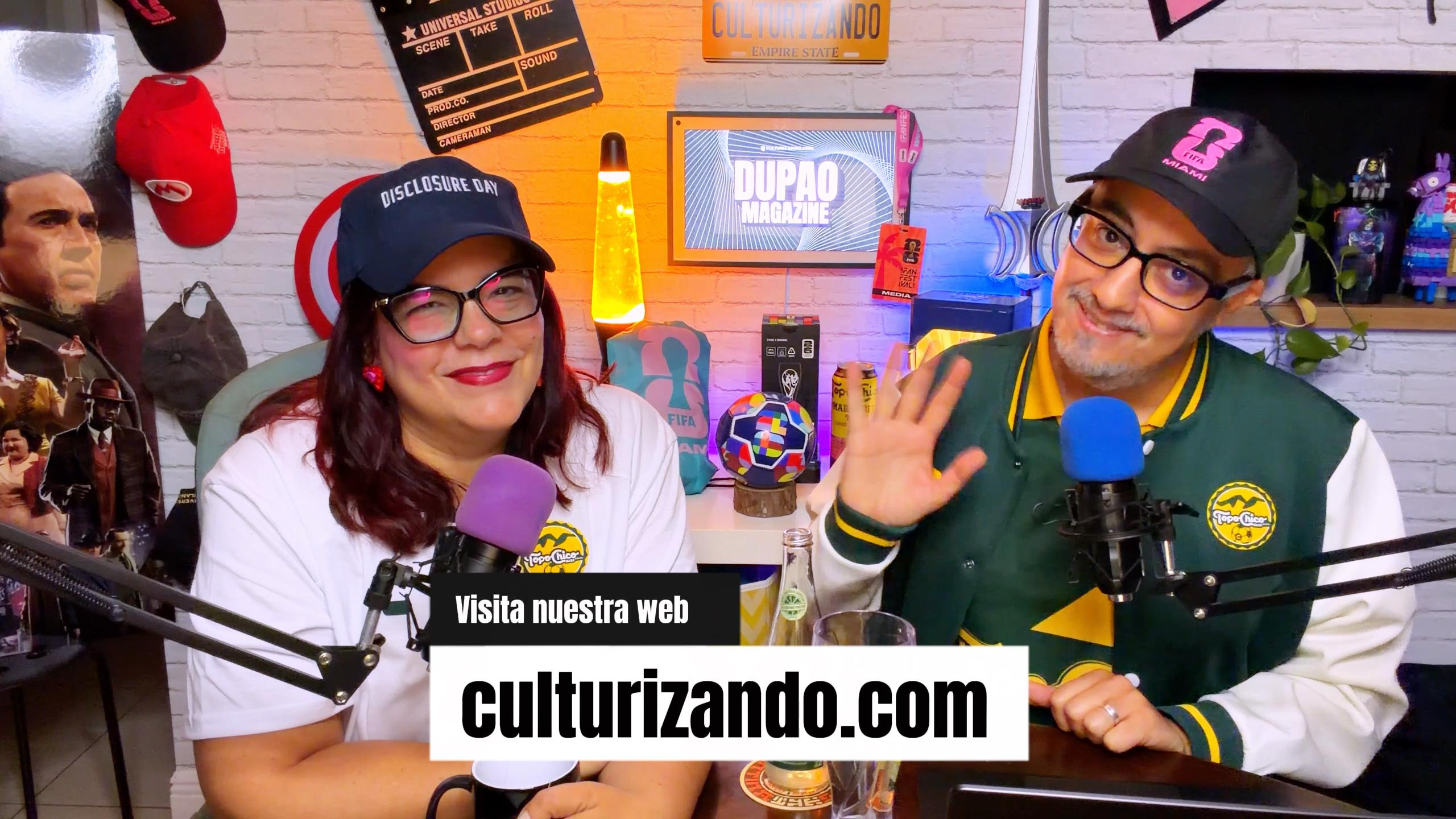 Two people sit at a table with microphones, in a colorful room with posters and bottles, smiling and waving.