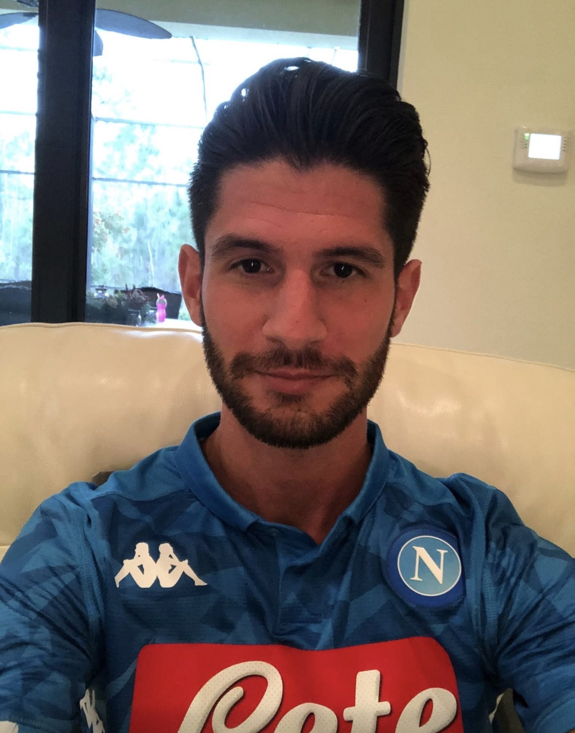 Young man with dark hair and beard wearing a blue sports jersey, sitting on a beige couch indoors.