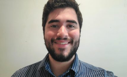 Young man with dark hair and beard smiling, wearing a striped shirt, against a plain light background.
