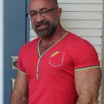 Man with glasses and beard wearing a red shirt with green and yellow trim, standing outdoors near a white wall.