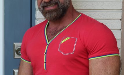 Man with glasses and beard wearing a red shirt with green and yellow trim, standing outdoors near a white wall.