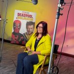 Woman with glasses and dark hair, wearing a yellow jacket, sitting on a chair in front of a Deadpool poster, studio lighting setup visible.