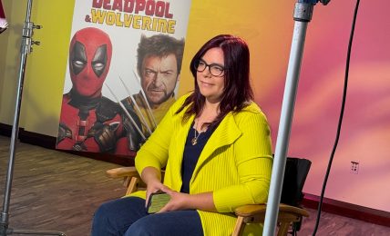 Woman with glasses and dark hair, wearing a yellow jacket, sitting on a chair in front of a Deadpool poster, studio lighting setup visible.