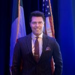 Man in a pinstripe suit standing in front of flags, smiling, with blue background.
