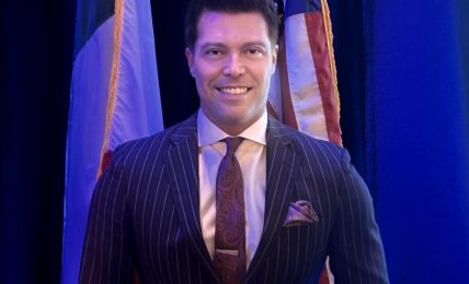 Man in a pinstripe suit standing in front of flags, smiling, with blue background.