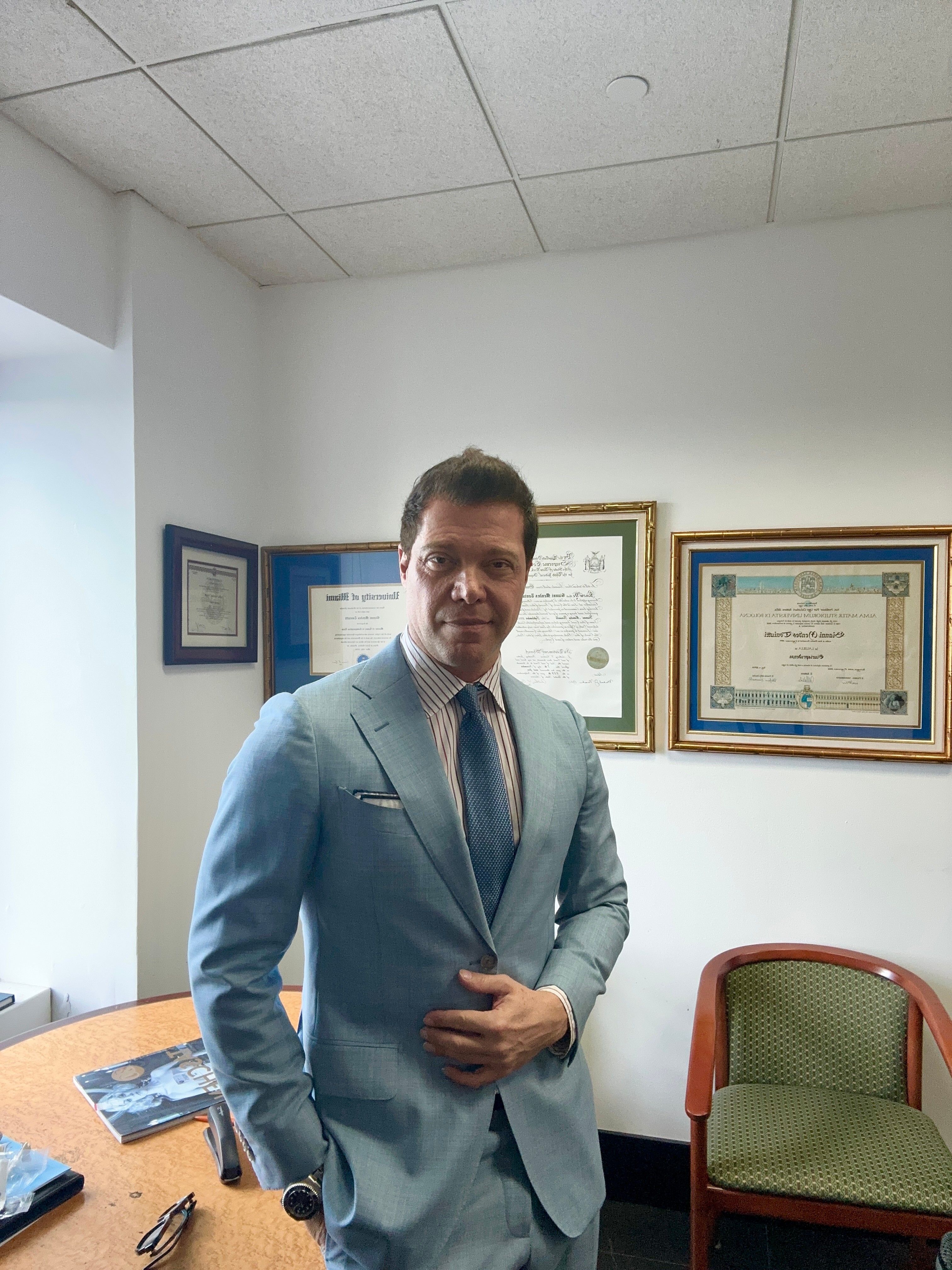 Man in a light blue suit standing in an office with framed certificates on the wall.