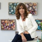 Woman with wavy brown hair in a white blazer and dark pants sitting in front of colorful artwork, surrounded by flowers.