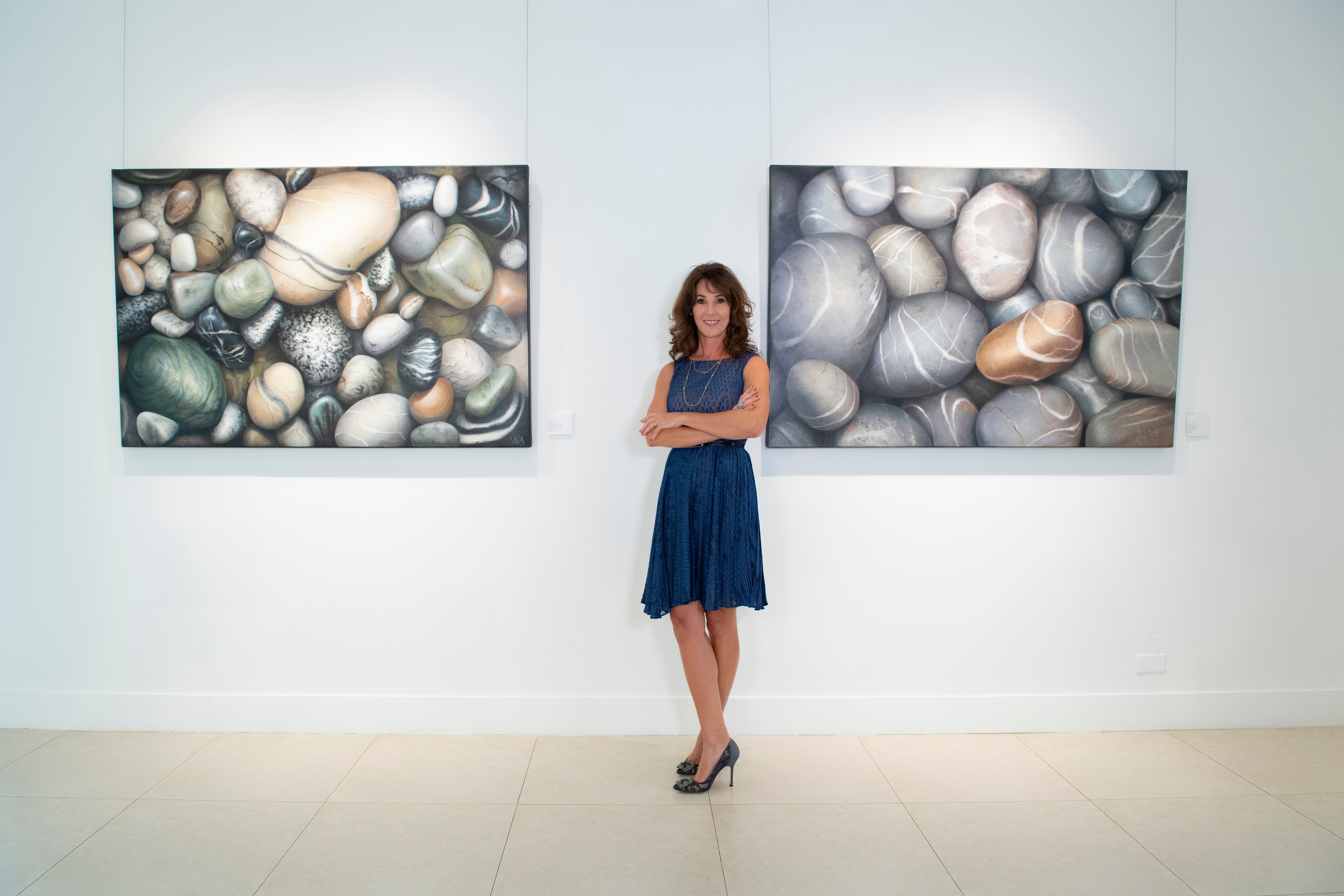 Woman in blue dress standing between two pebble artwork displays in gallery, arms crossed, white wall background.