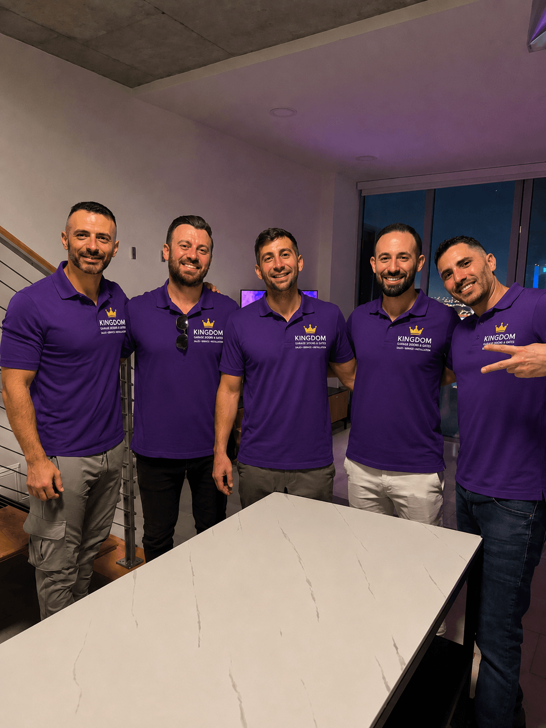 Five men in purple shirts standing indoors, smiling, with a table in front of them.