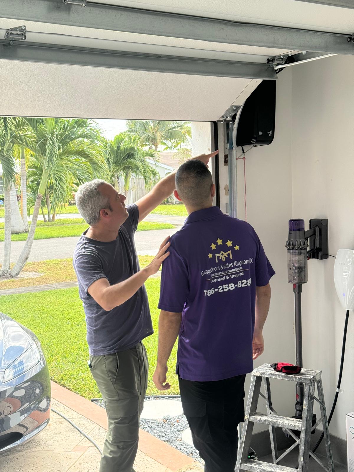 Two men working on a garage door opener, one pointing at the device, with trees and a car outside.