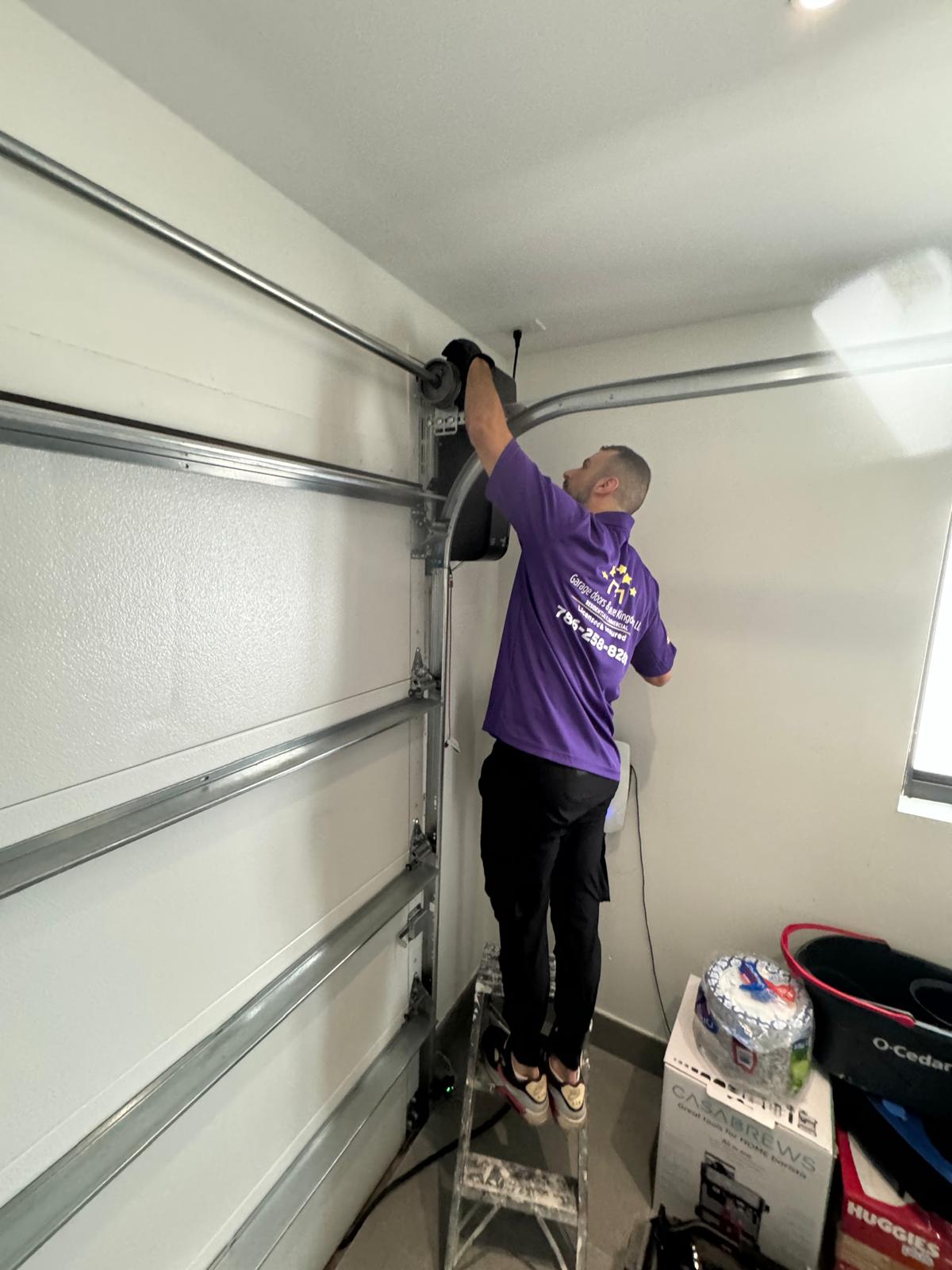Person standing on a ladder working on garage door mechanism, wearing a purple shirt and black pants, in a garage.