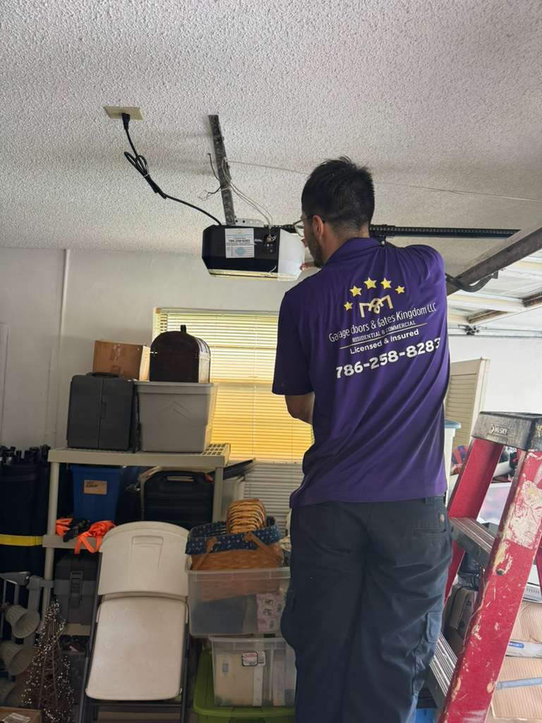 Man working on ceiling with tools, wearing a purple shirt with stars and text, in a cluttered room.