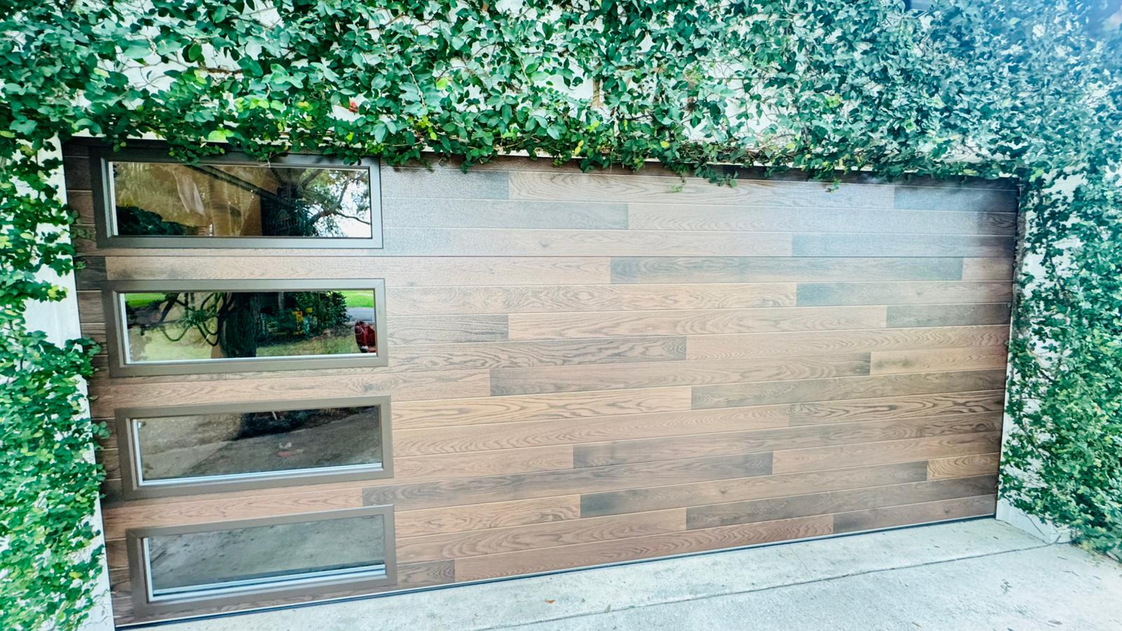 Wooden garage door with three small windows on the left, surrounded by green foliage, and a concrete driveway below.