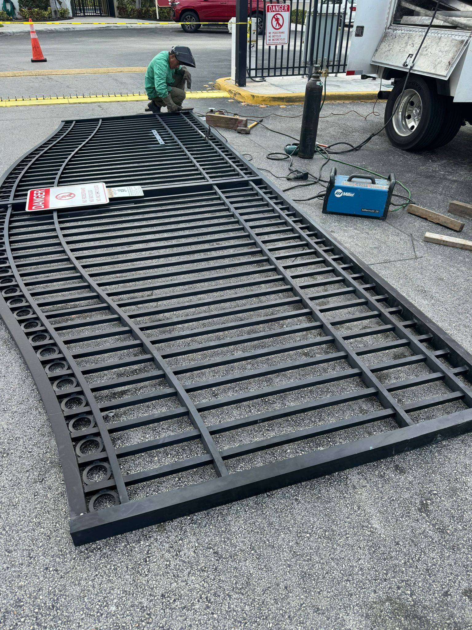 Worker installing a curved metal gate on a street, with tools and equipment nearby, and a truck parked beside.