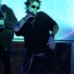 Person with spiky hair, sunglasses, and jewelry poses in a neon-lit environment, wearing a black outfit with chains.