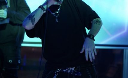 Person with spiky hair, sunglasses, and jewelry poses in a neon-lit environment, wearing a black outfit with chains.