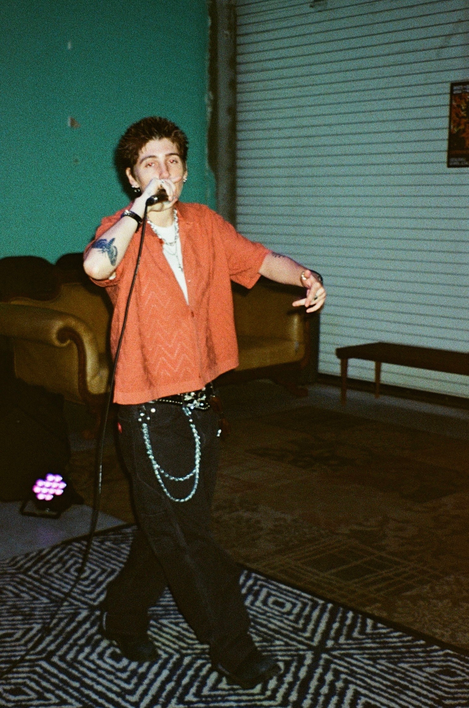 Person singing into a microphone on stage with a green wall and closed window behind, wearing a red shirt and black pants.