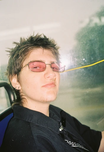 Person with spiky hair wearing pink-tinted glasses, sitting in a vehicle, looking at the camera.