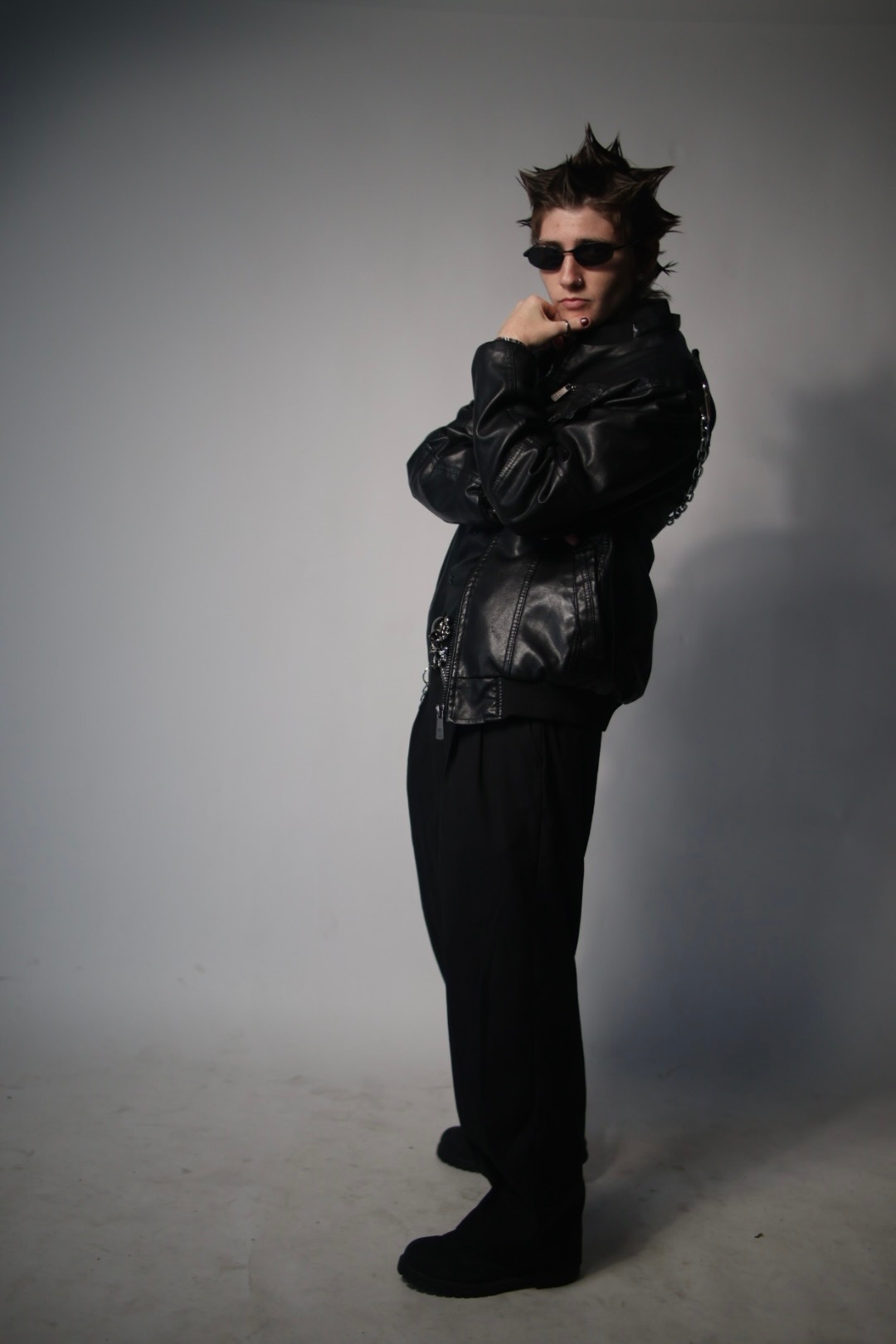 Person with spiky hair wearing sunglasses and a black leather jacket, standing with arms crossed against a plain background.