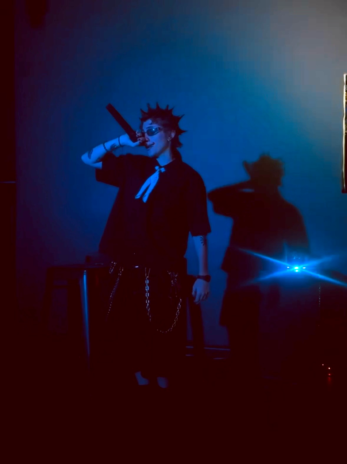 Person with spiked hair and sunglasses holding a microphone, shadow cast on blue-lit background.