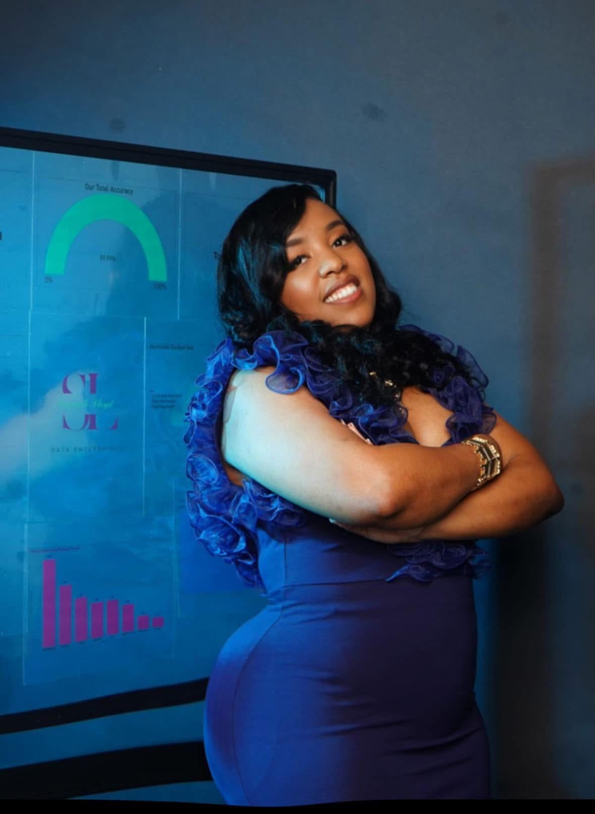 Woman with long curly hair smiling, arms crossed, standing in front of a screen displaying colorful graphs and charts.