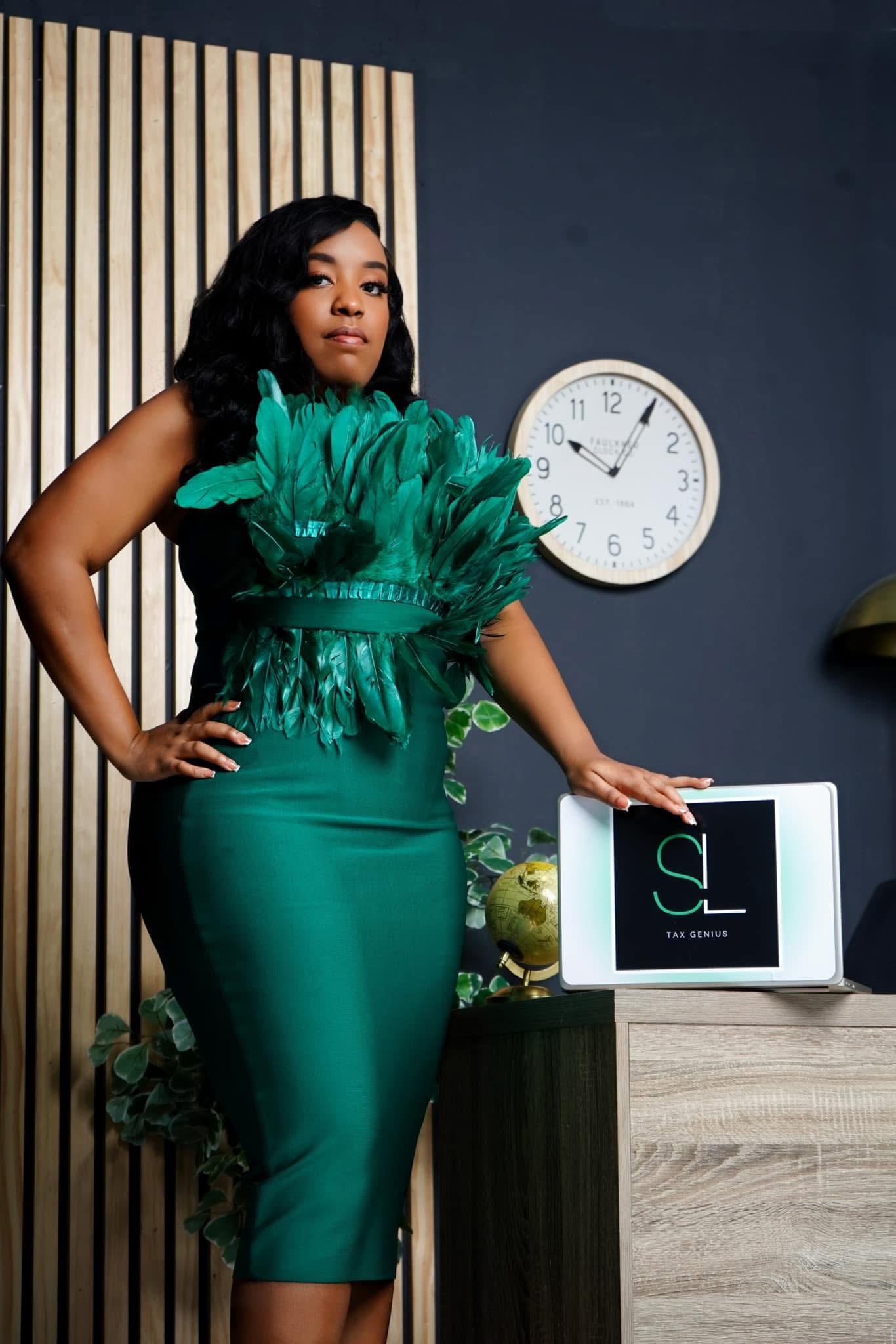 Woman in green dress standing next to a desk with a computer monitor, in a room with a clock on the wall.