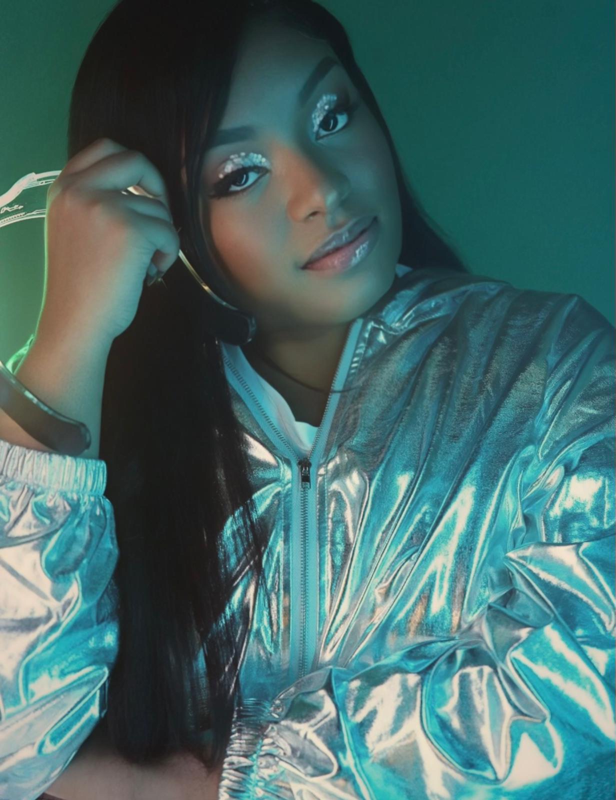 Young woman with long dark hair, wearing shiny silver jacket, smiling, with makeup and earrings, in a dimly lit setting.
