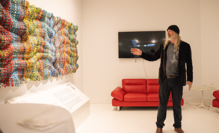 Person with long hair, black hat, and dark clothing points at a wall-mounted TV in a white gallery room with colorful art installation.