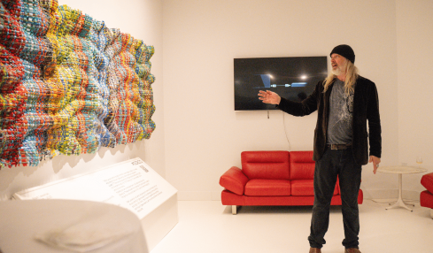 Person with long hair, black hat, and dark clothing points at a wall-mounted TV in a white gallery room with colorful art installation.