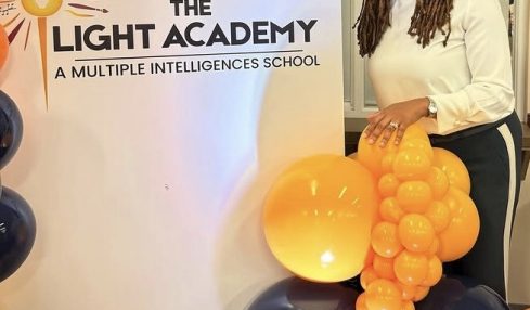 Woman standing next to a sign for The Light Academy, holding a cluster of yellow and black balloons, smiling.