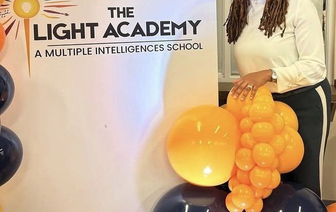 Woman standing next to a sign for The Light Academy, holding a cluster of yellow and black balloons, smiling.