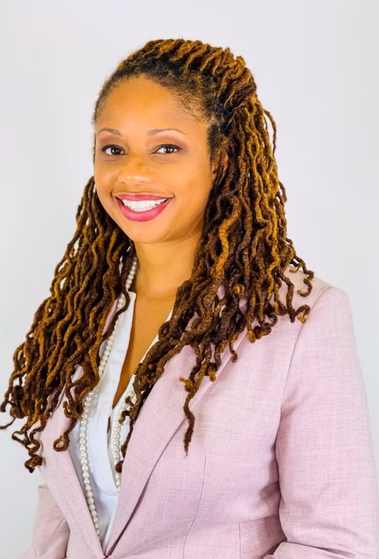 Portrait of a smiling woman with long, styled dreadlocks, wearing a light pink blazer and white top, against a plain background.