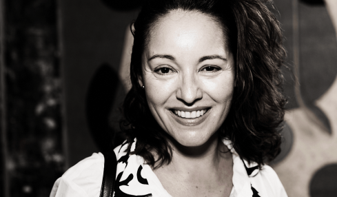 Smiling woman with curly hair wearing a patterned blouse, standing in front of abstract artwork.