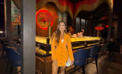 Woman in yellow blazer dress standing in a dimly lit restaurant with colorful decor and seating.