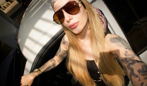 Woman with long blonde hair wearing sunglasses, a headscarf, and tattoos, taking a selfie indoors.