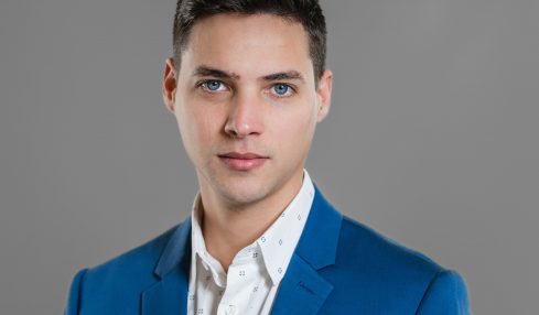 Young man with dark hair and blue eyes wearing a blue blazer and white shirt against a gray background.