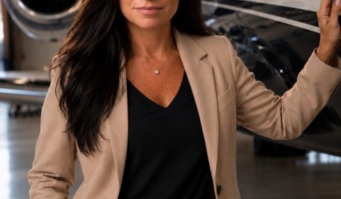 Woman in beige suit standing next to private jet in hangar, looking at camera.