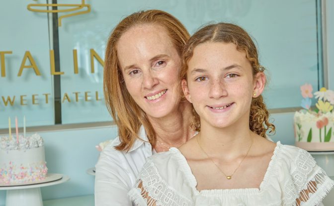 Two females smiling, one adult and one child, in front of a decorated cake and pastel background.